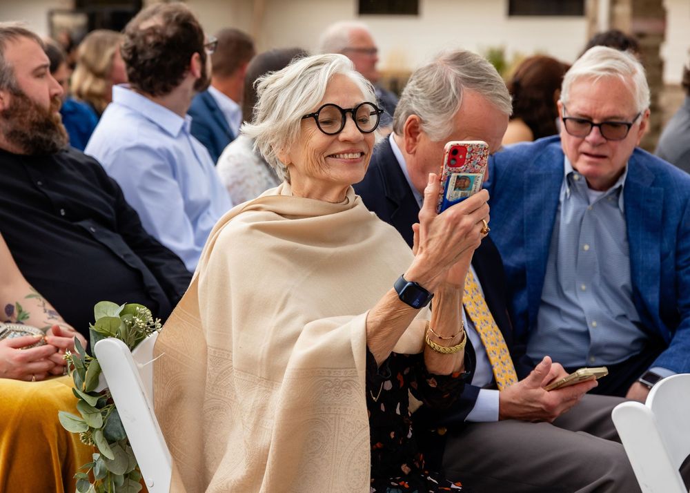 Elderly woman with white hair and glasses smiling while taking a photo with her phone at a formal gathering.