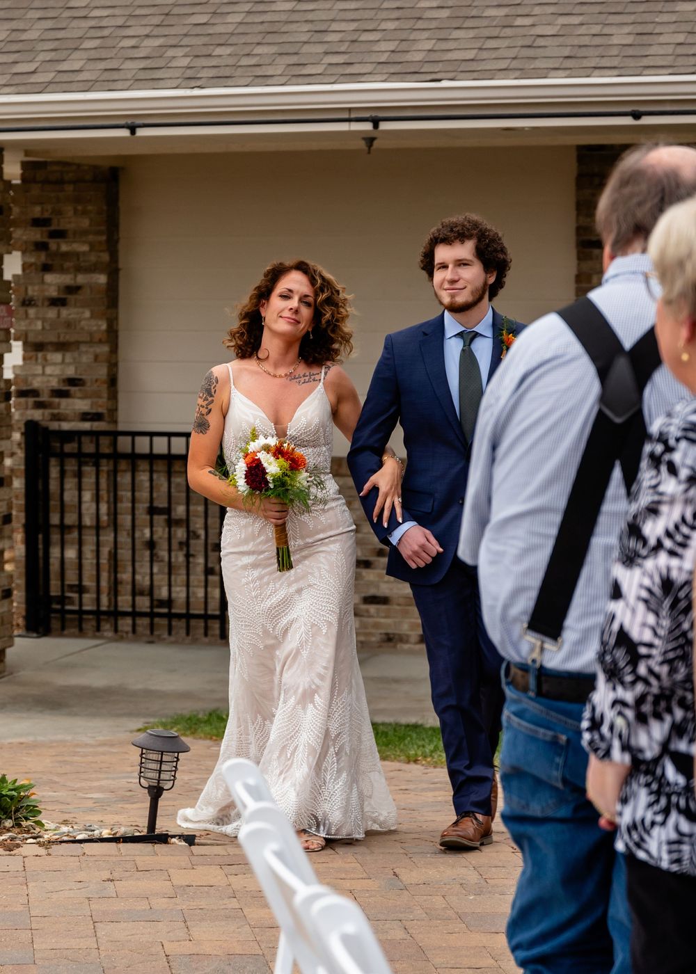 Bride in white lace gown holding colorful bouquet walks with son in navy suit at outdoor wedding ceremony at a Venue on the Ridge in Elkhorn