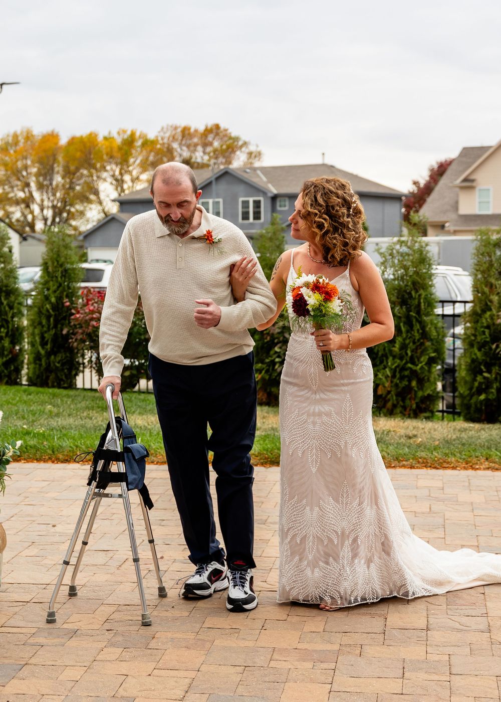 Bride in lace gown walks beside her dad using a walker on a brick patio during an outdoor wedding ceremony at a Venue on the Ridge