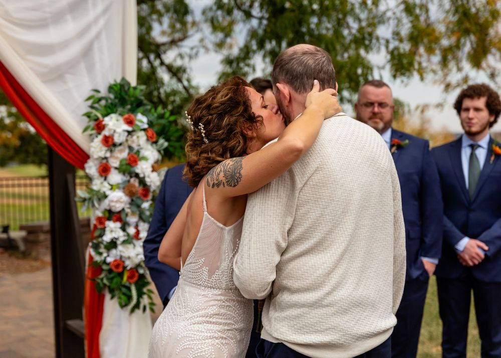 Bride and groom share first kiss at outdoor wedding ceremony with floral arch and guests in background at a Venue on the Ridge in Elkhorn NE