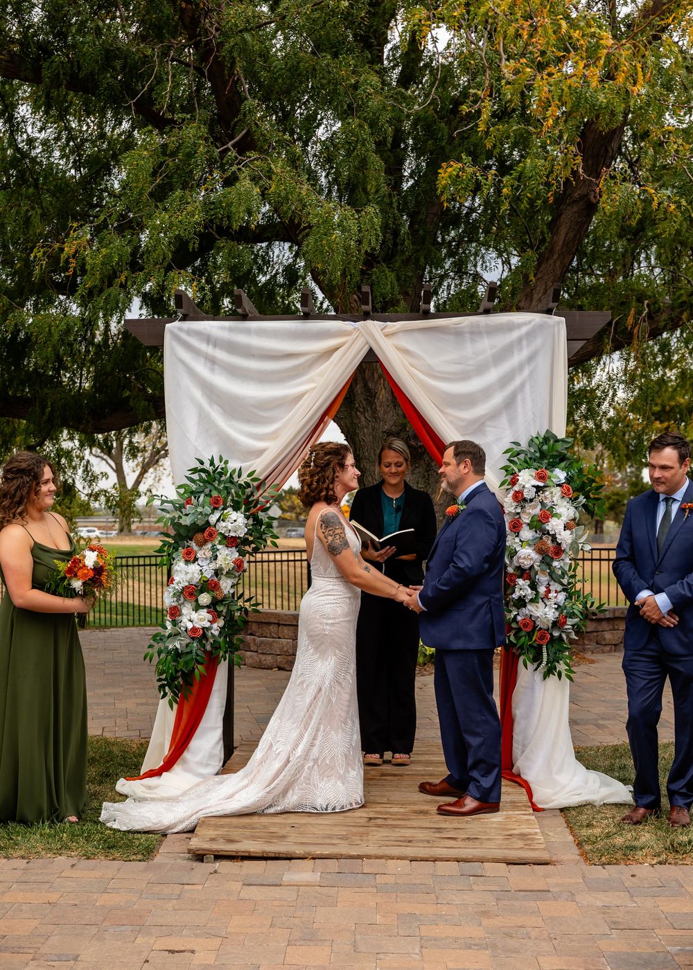 Bride and groom exchange vows at an outdoor wedding altar adorned with white draping and red and white floral arrangements