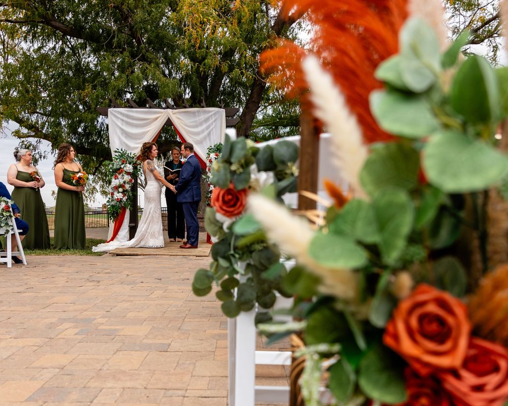 Outdoor wedding ceremony with couple at floral arch, flanked by guests, with orange and green floral arrangement in foreground in Elkhorn