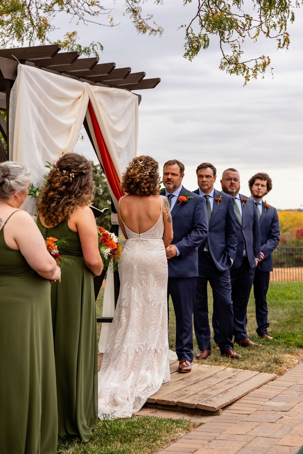 Outdoor wedding ceremony with bride in white lace gown facing groomsmen in navy suits under a draped pergola at a Venue on the Ridge