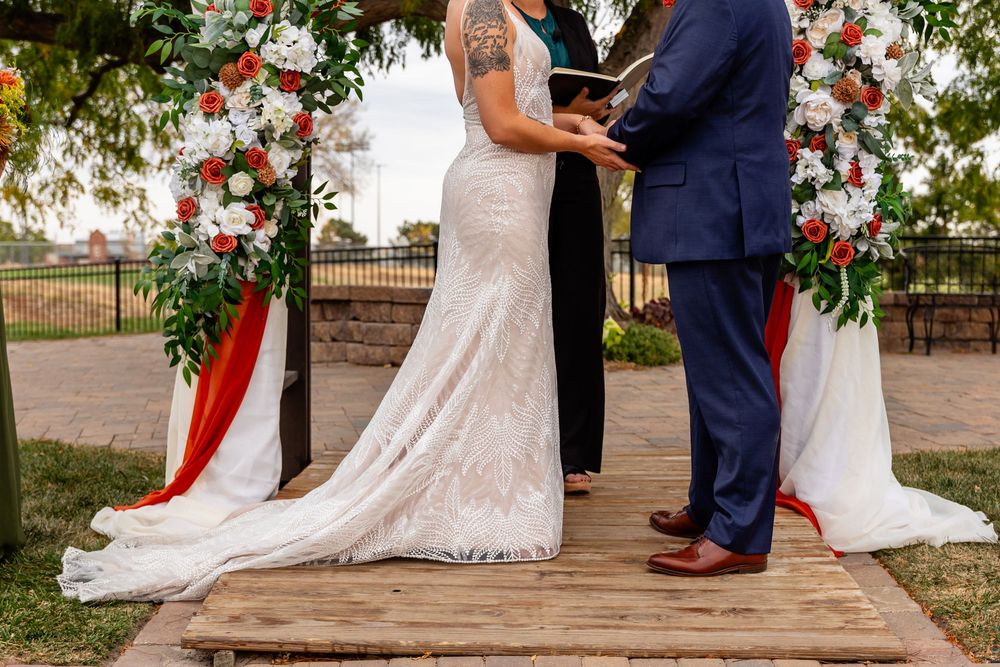 Bride and groom exchange vows at outdoor wedding ceremony with red and white floral arch on wooden platform.