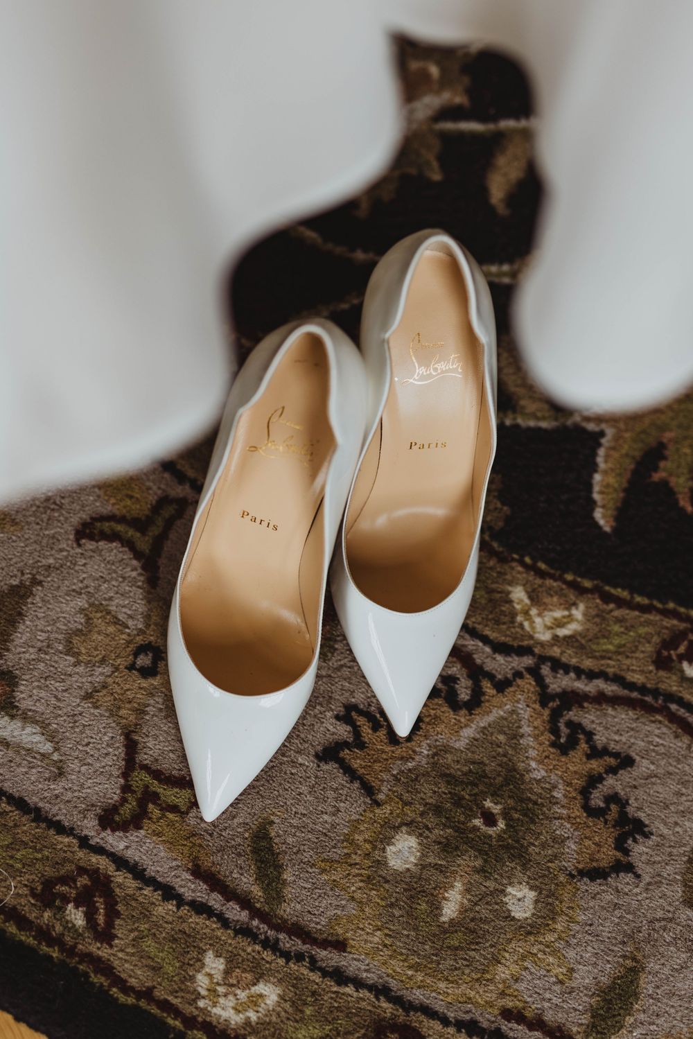 White Christian Louboutin pointed-toe heels displayed on a floral patterned rug at a wedding.