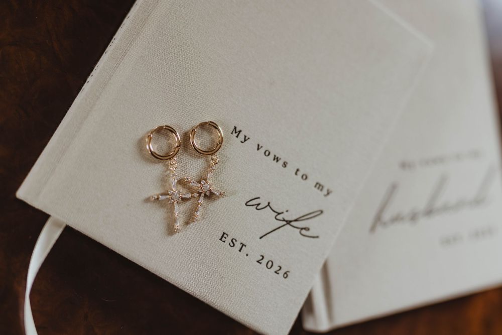 Gold hoop earrings rest on wedding vow books labeled My Vows to My Wife and Husband EST. 2026.