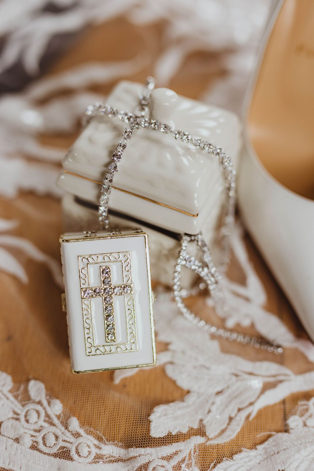 Bridal accessories flat lay featuring a crystal cross locket necklace, diamond ring box, and white wedding heel on lace.