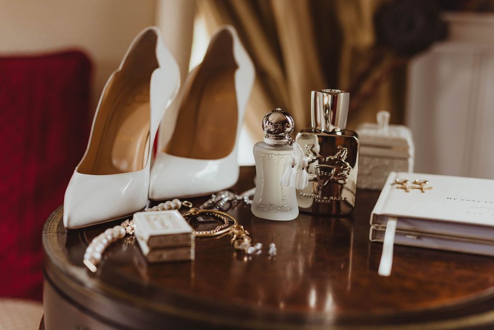 White Louboutin heels, pearl jewelry, perfume bottles, and wedding accessories arranged on a wooden table.