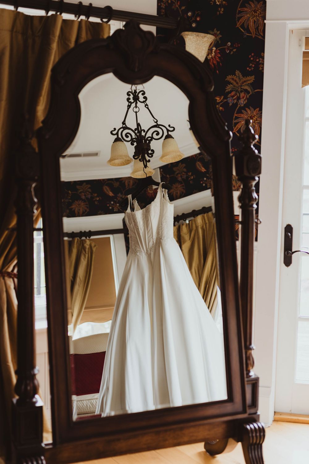 White wedding gown hanging from a chandelier reflected in an ornate dark wood floor mirror.