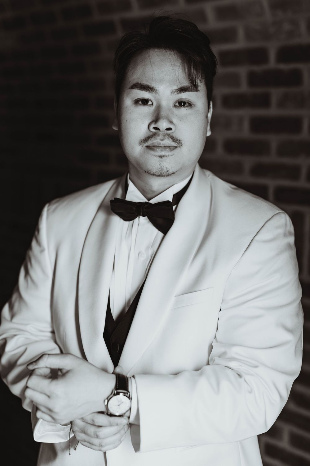 Groom in white tuxedo jacket and black bow tie poses confidently in black and white portrait photo.