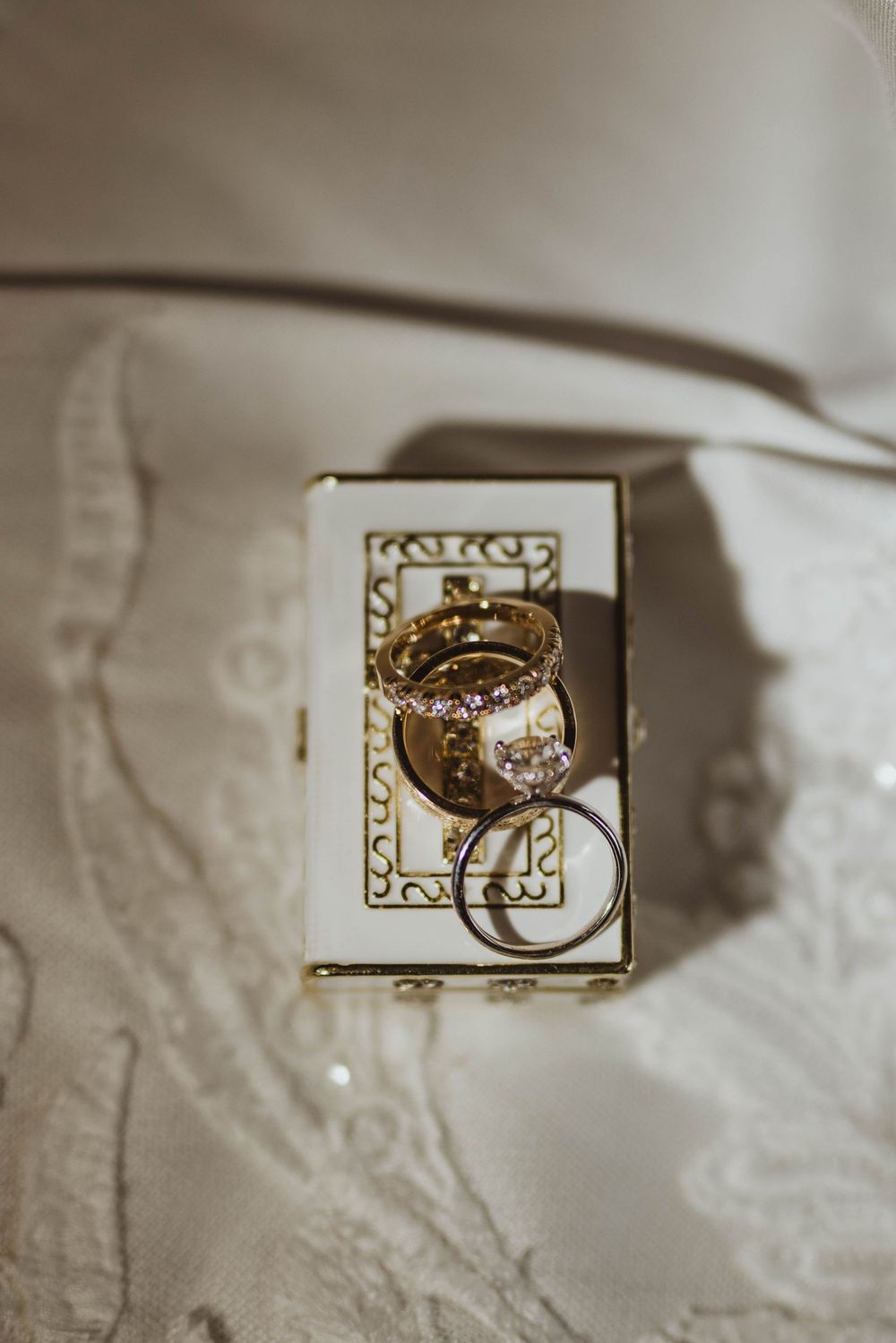 Wedding rings and gold bands displayed on an ornate white and gold jewelry box atop lace fabric.