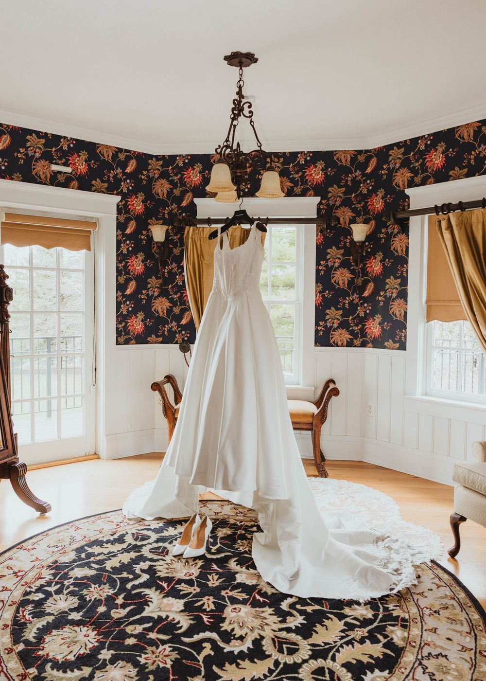 Elegant white wedding gown with long train hanging from chandelier in a room with dark floral wallpaper.
