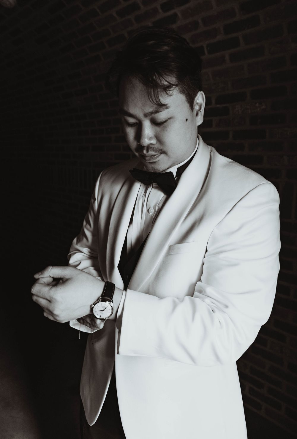 Groom in white tuxedo jacket adjusting his watch in a black and white portrait photo.