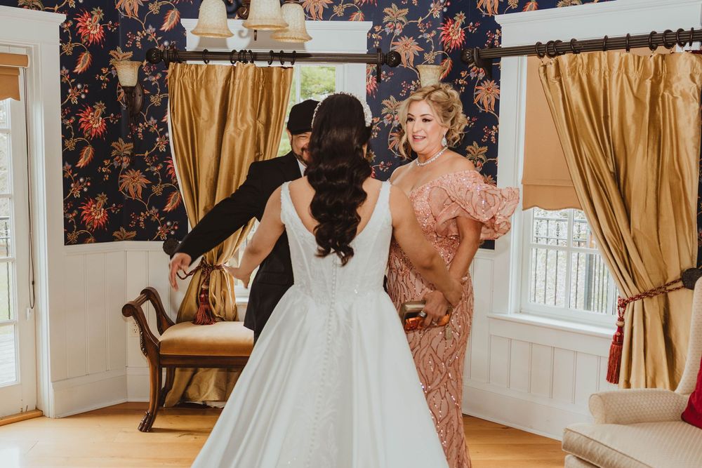 Bride in white ball gown sharing a tender moment with family members in a floral wallpapered room.