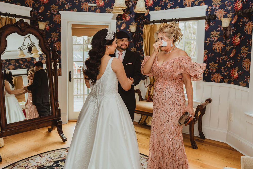 Bride in white gown shares emotional moment with father and tearful mother in floral wallpapered room.