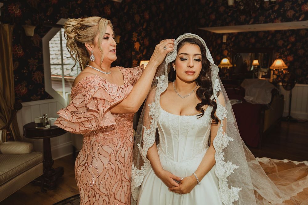 Mother helping bride with lace veil in floral wallpapered bridal suite before wedding.