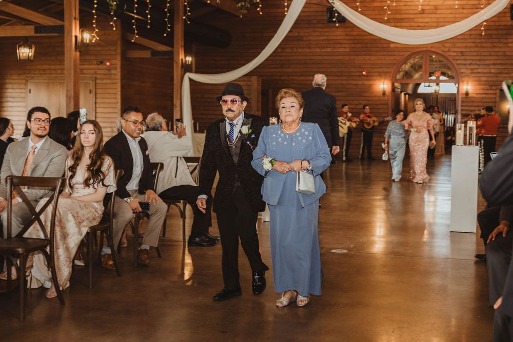 Wedding guests walk down the aisle in a rustic barn ceremony venue with warm string lights overhead.