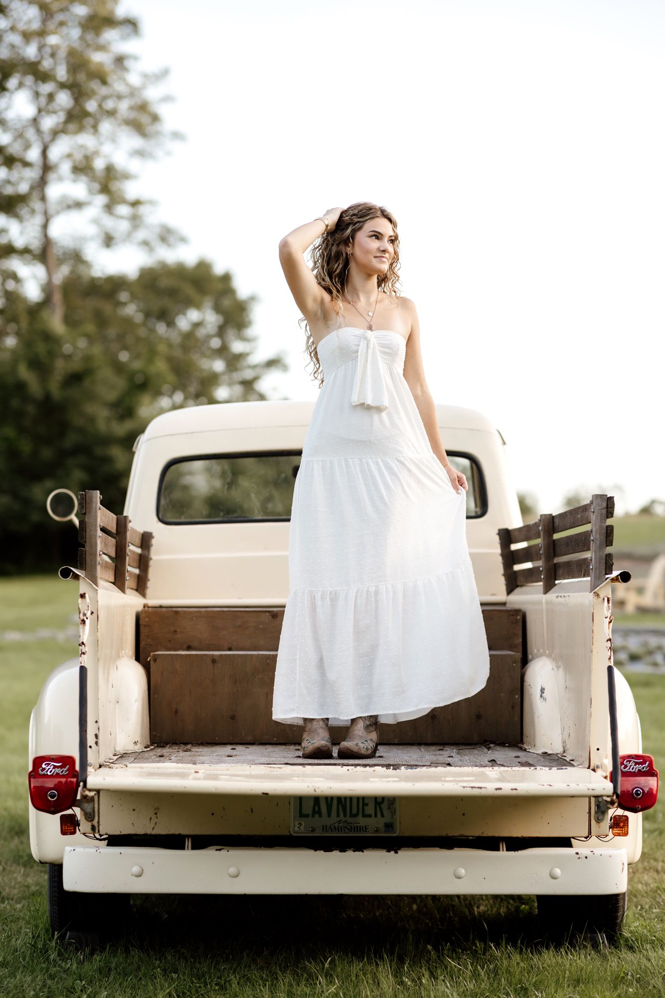 Senior photos of a young lady wearing off-shoulder white maxi dress posing in bed of pick up truck at Pumpkin Blossom Warner, New Hampshire