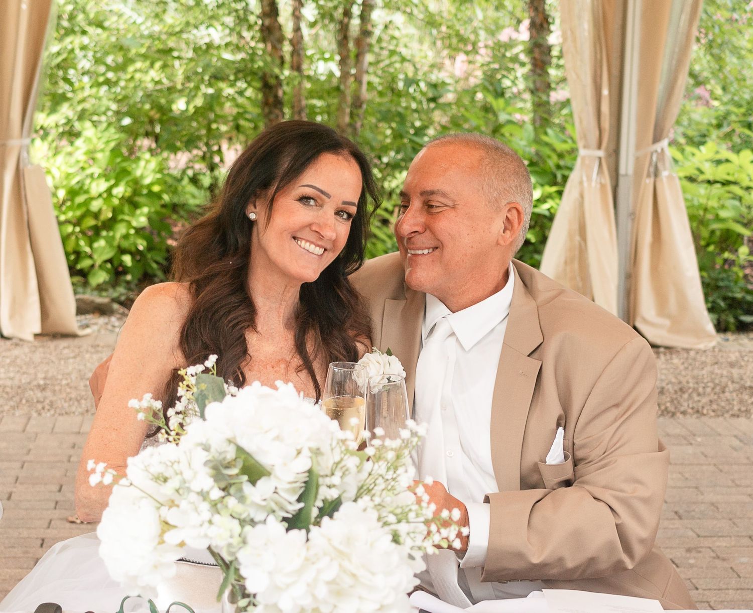 Happy couple smiling at each other at wedding reception with white floral centerpiece and elegant outdoor venue.