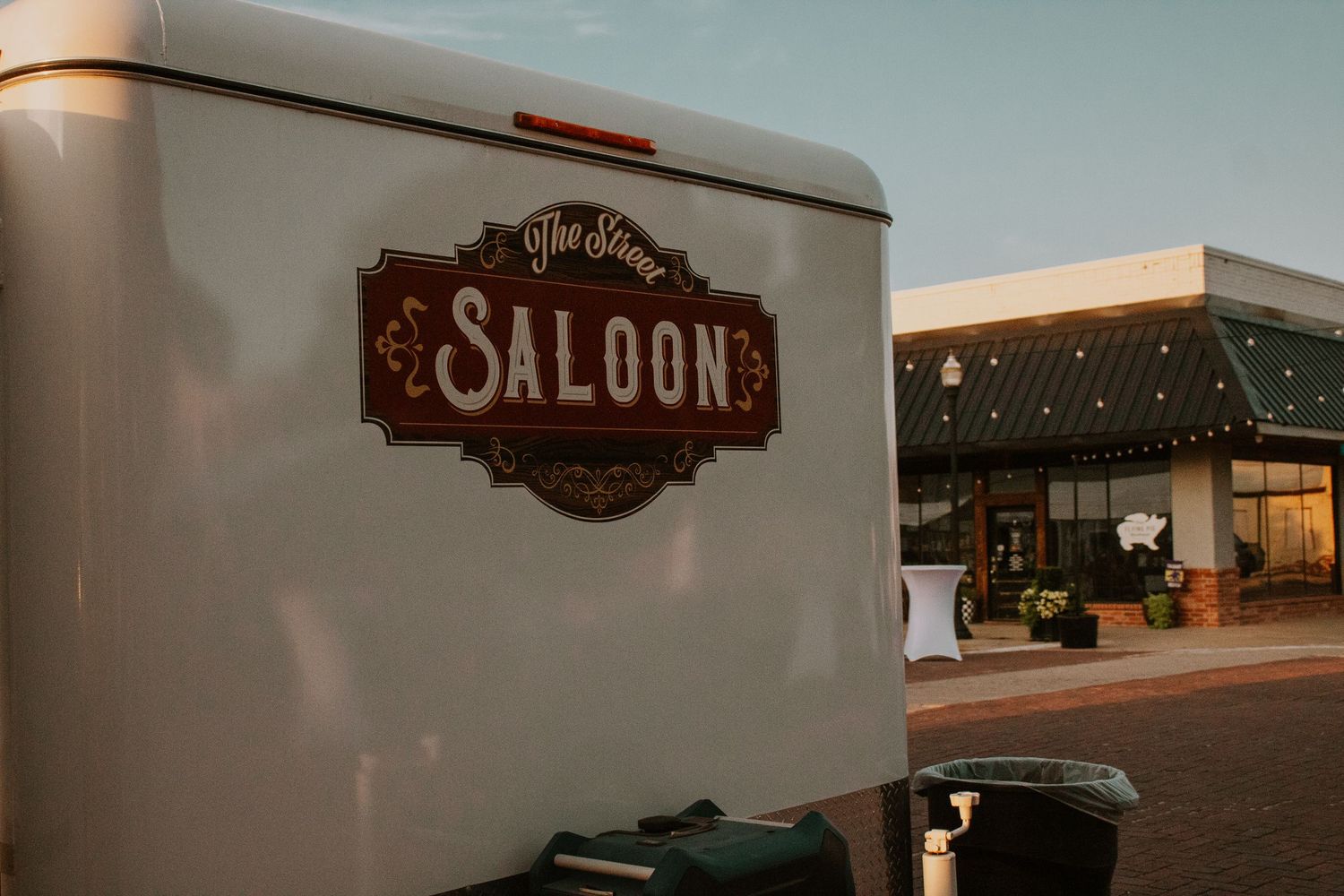 A vintage white trailer with a decorative Saloon sign parked outside a retail storefront at dusk.
