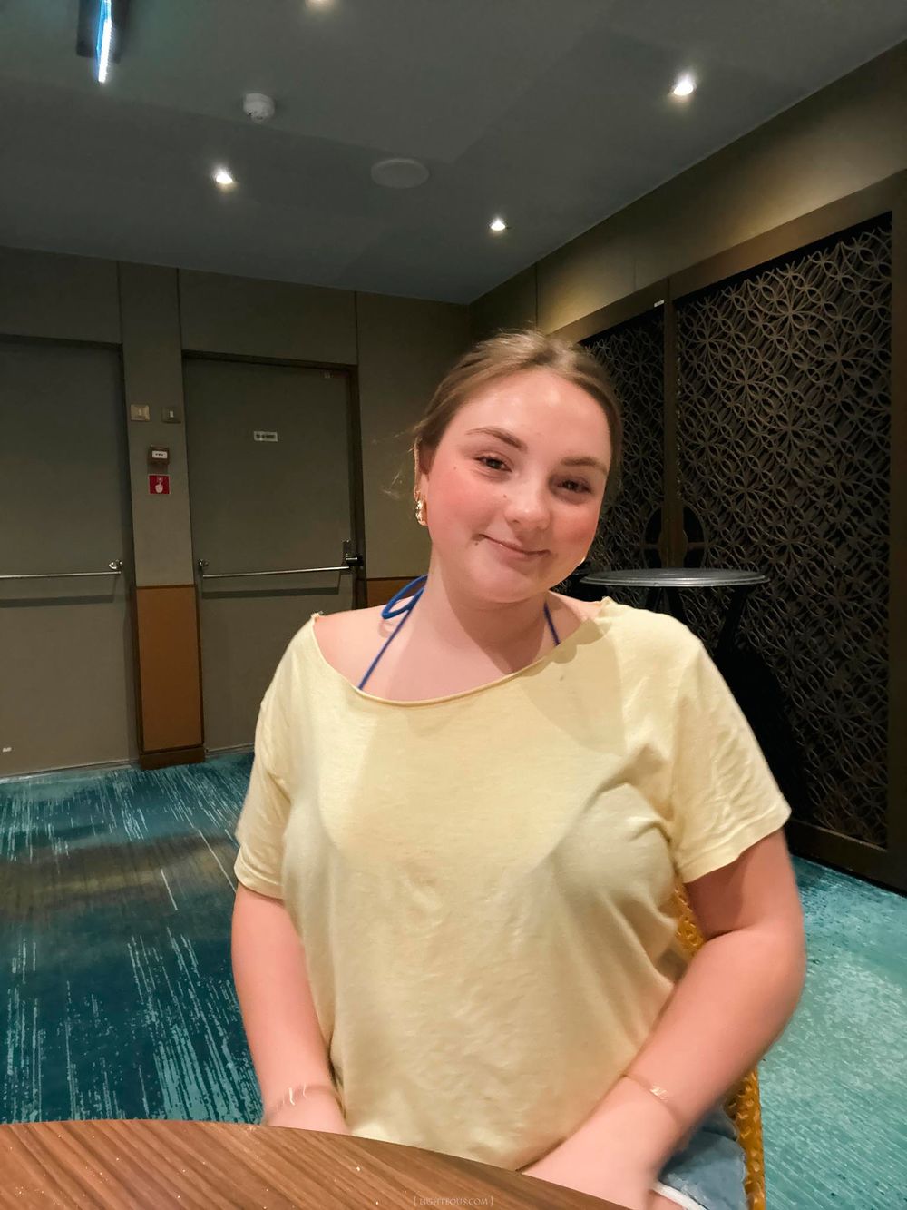 Young girl in yellow t-shirt smiling at camera inside modern hotel lobby with teal carpet.