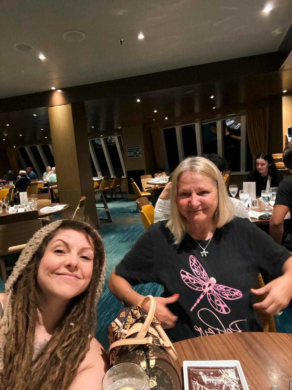 Two women smiling at a restaurant table, one with dreadlocks and one in a dragonfly t-shirt, dining in a warmly lit venue.