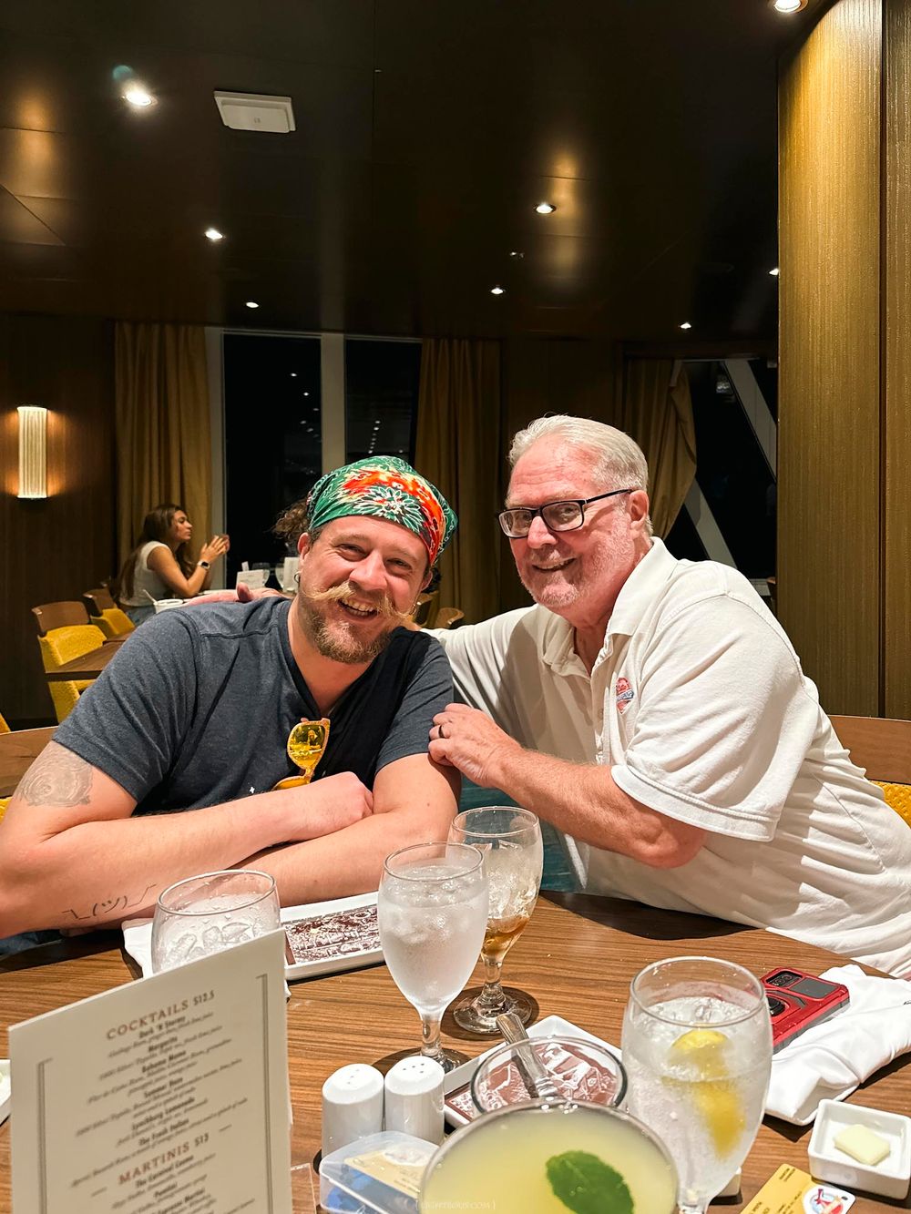 Two men smiling and posing together at a restaurant table with cocktail menus and drinks.