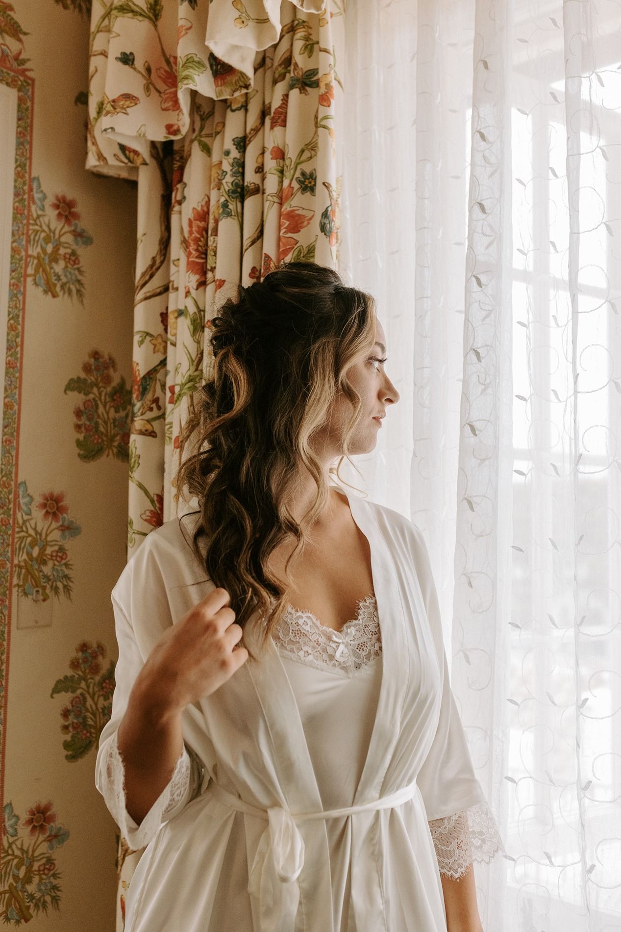 Bride in white lace robe gazing out window with floral curtains on her wedding morning at Darlington House.