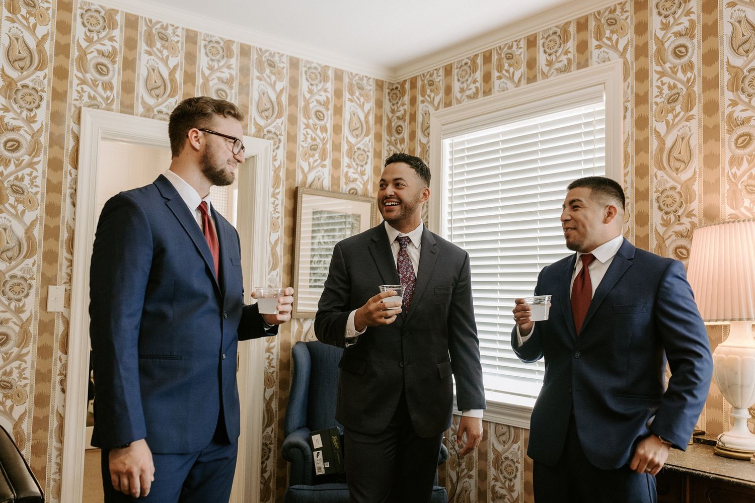 A groom and two groomsmen in suits socializing and holding drinks in a decorative wallpapered room before a wedding.