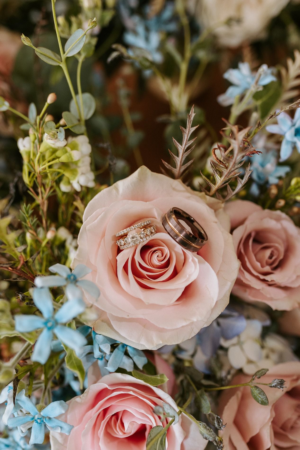 Two gold wedding rings rest inside a pink rose surrounded by a bouquet of blush roses and delicate blue flowers.