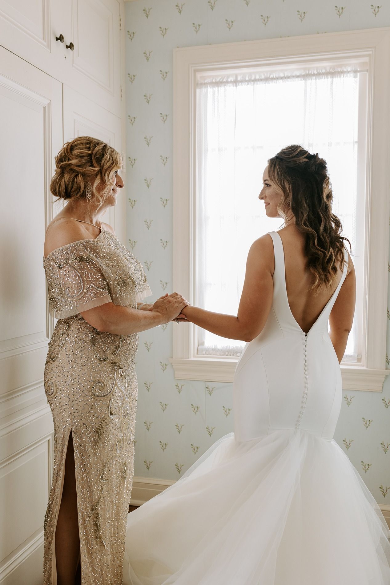 Bride in backless gown holds hands with mother in beaded dress by window in floral wallpaper room at Darlington House.