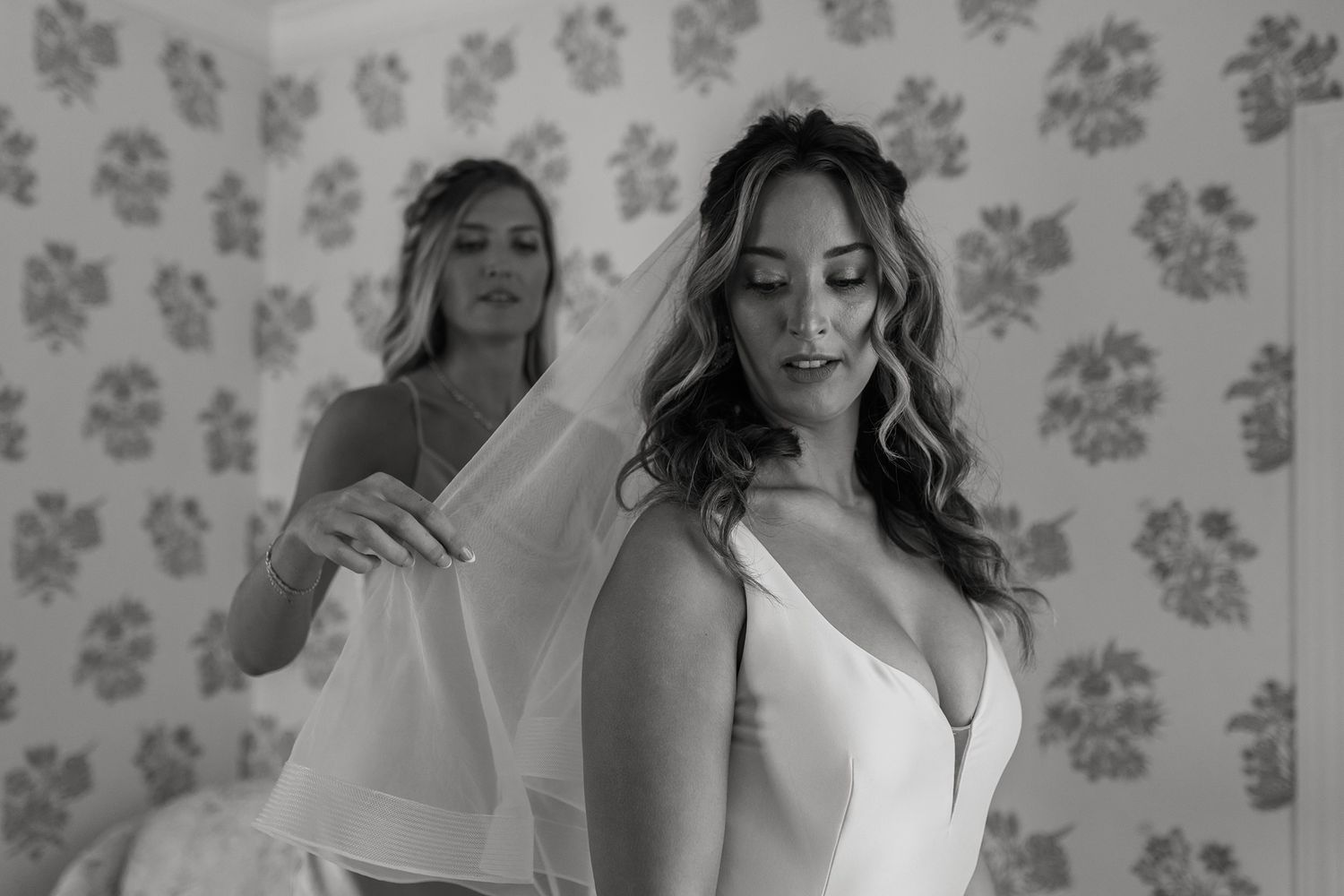 Black and white photo of a bride in a white gown being helped by a bridesmaid in a floral wallpaper room at Darlington House.