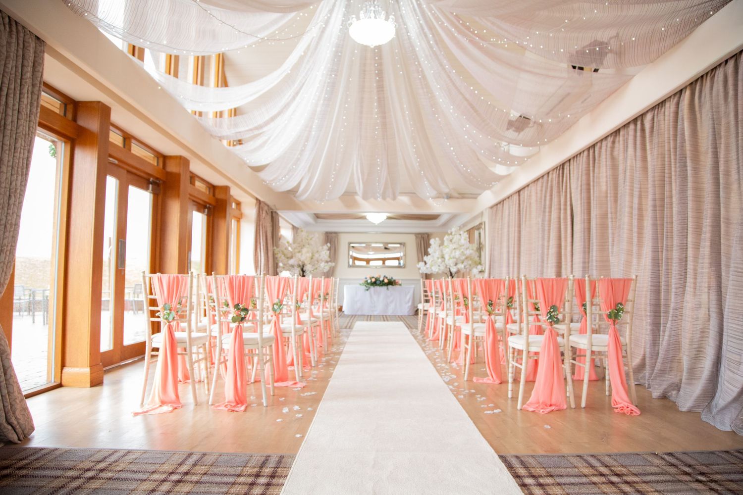 Elegant wedding ceremony venue with pink satin chair sashes, white aisle runner, and draped ceiling decor.
