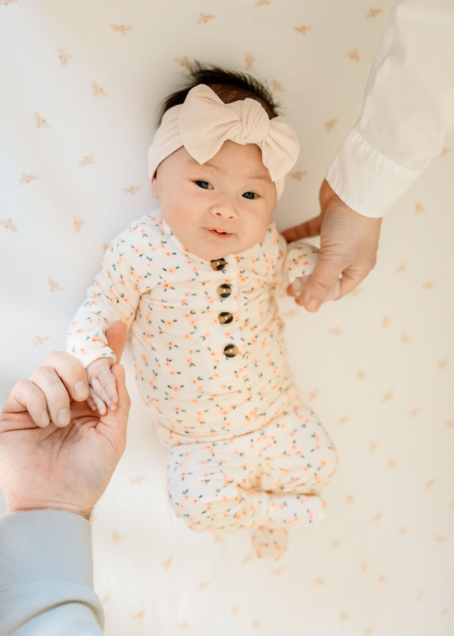 Newborn baby in floral onesie and bow headband lying in crib, held by two adults hands.