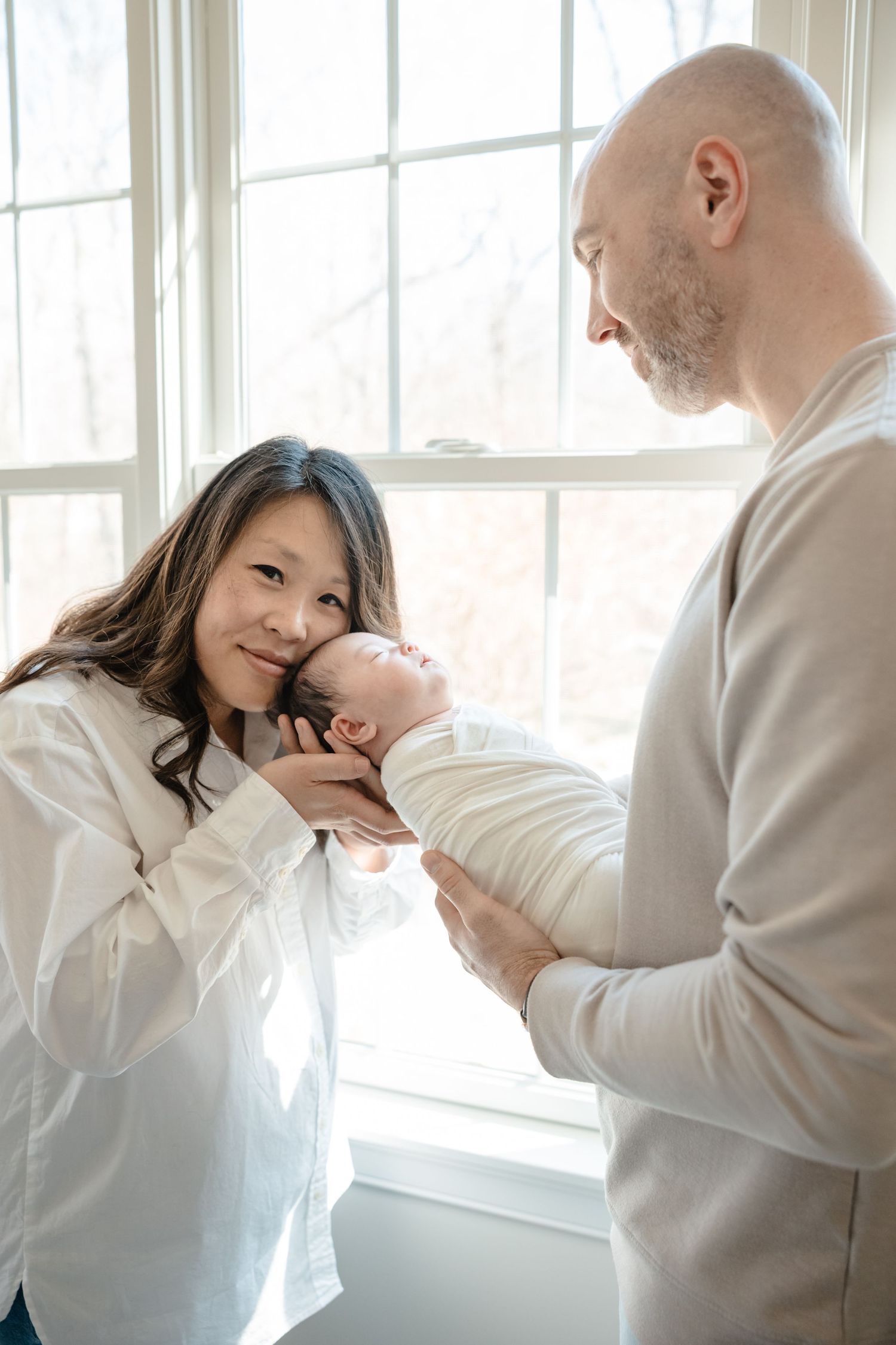 Parents lovingly holding their swaddled newborn baby near a bright window in a warm home setting.