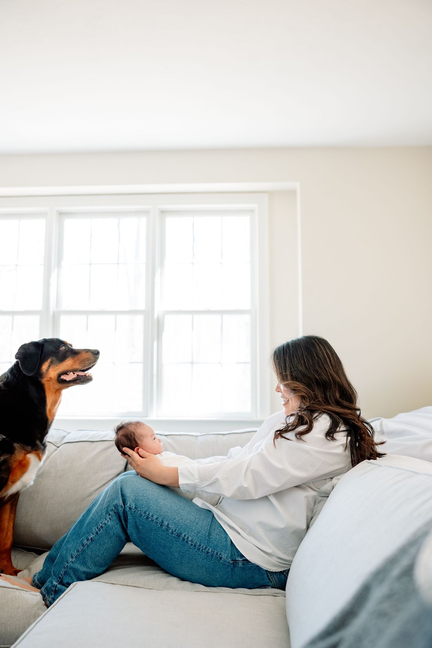 Mother sitting on couch holding newborn baby while family dog watches attentively near window.