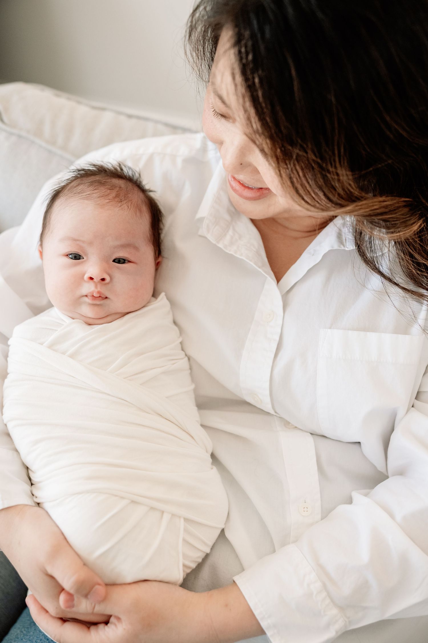 Mother in white shirt tenderly holding swaddled newborn baby with eyes open looking at camera.