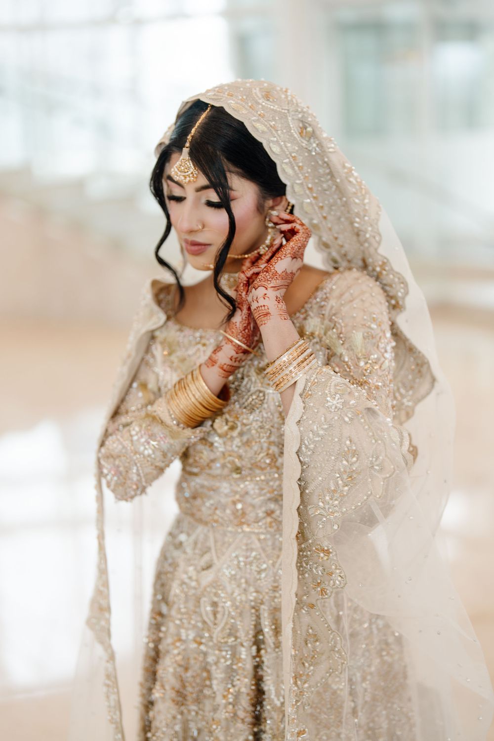 South Asian bride in ivory gold embroidered lehenga adjusts her dupatta veil with henna-adorned hands.