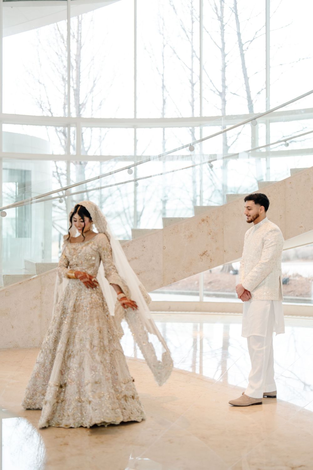 South Asian bride and groom share first look moment in ivory embroidered wedding attire on marble staircase.