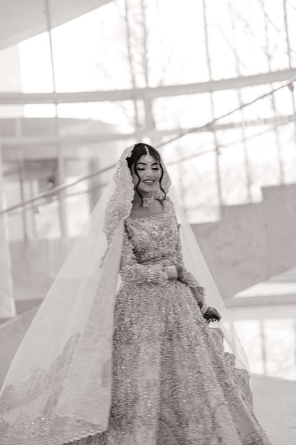 Black and white image of smiling South Asian bride in embroidered lehenga and flowing veil on staircase.