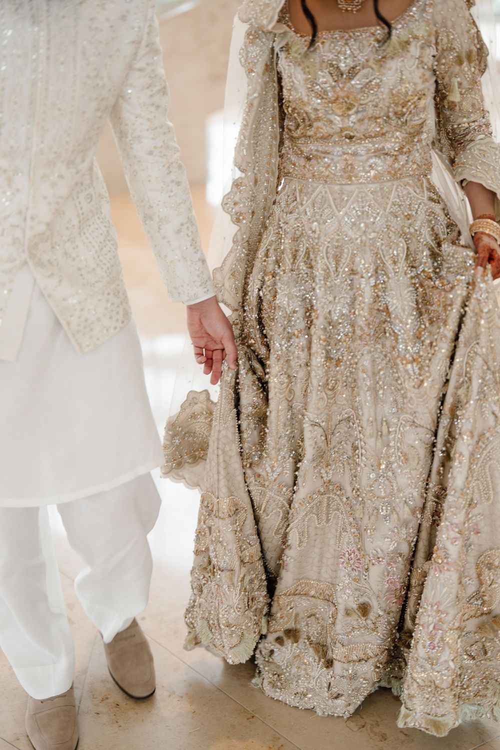 Detail shot of South Asian couple holding hands in matching ivory gold heavily embroidered wedding outfits.