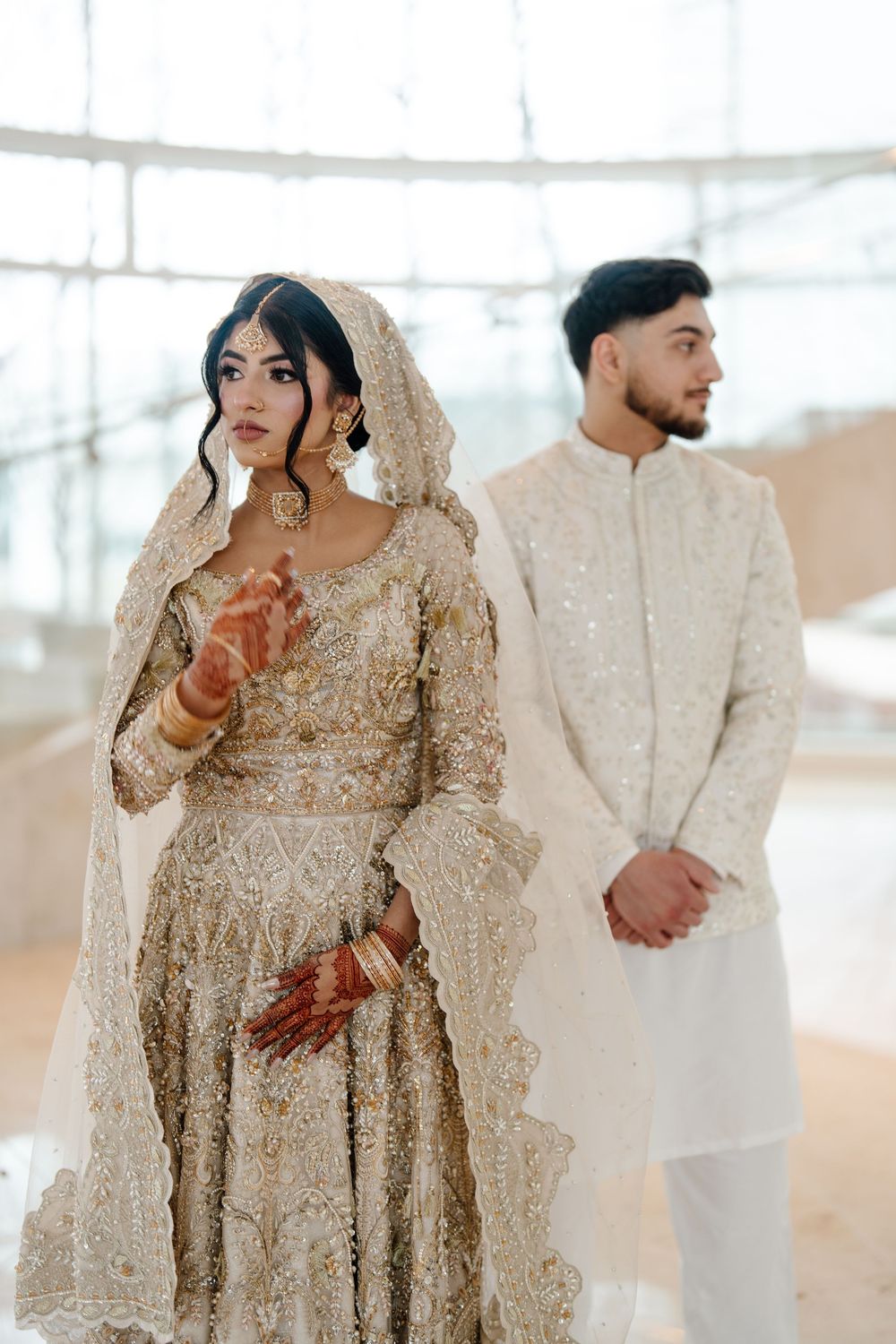 South Asian bride in gold embroidered lehenga with dupatta veil poses with groom in ivory sherwani.