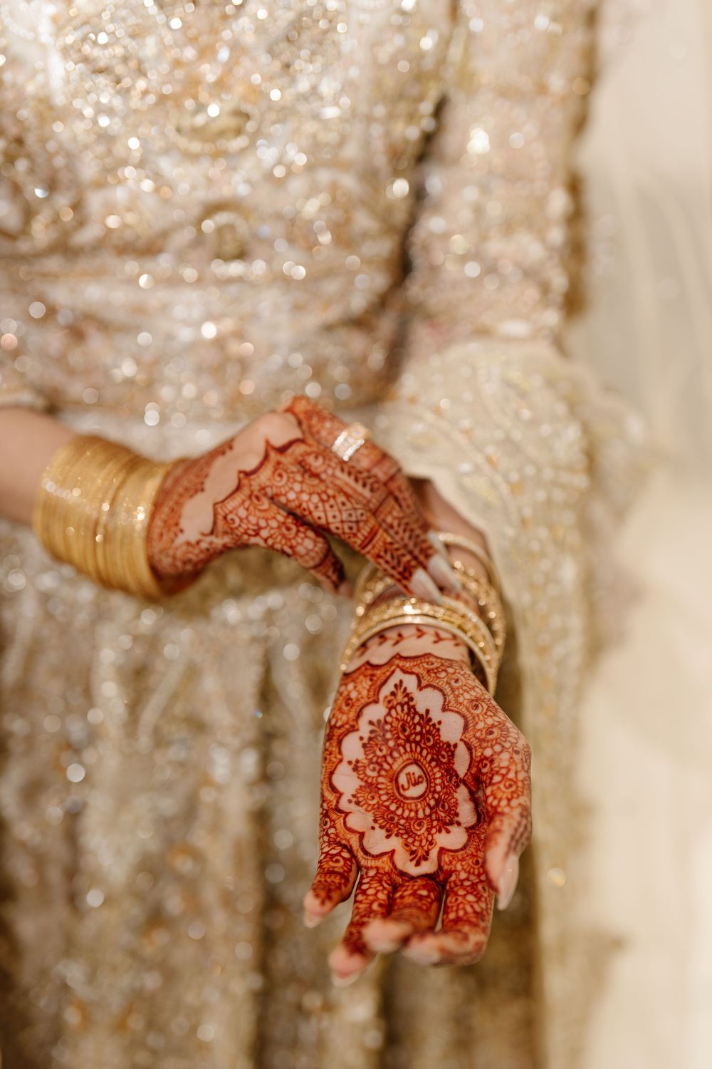 Close-up of bridal hands adorned with intricate red mehndi henna and gold bangles on embroidered lehenga.