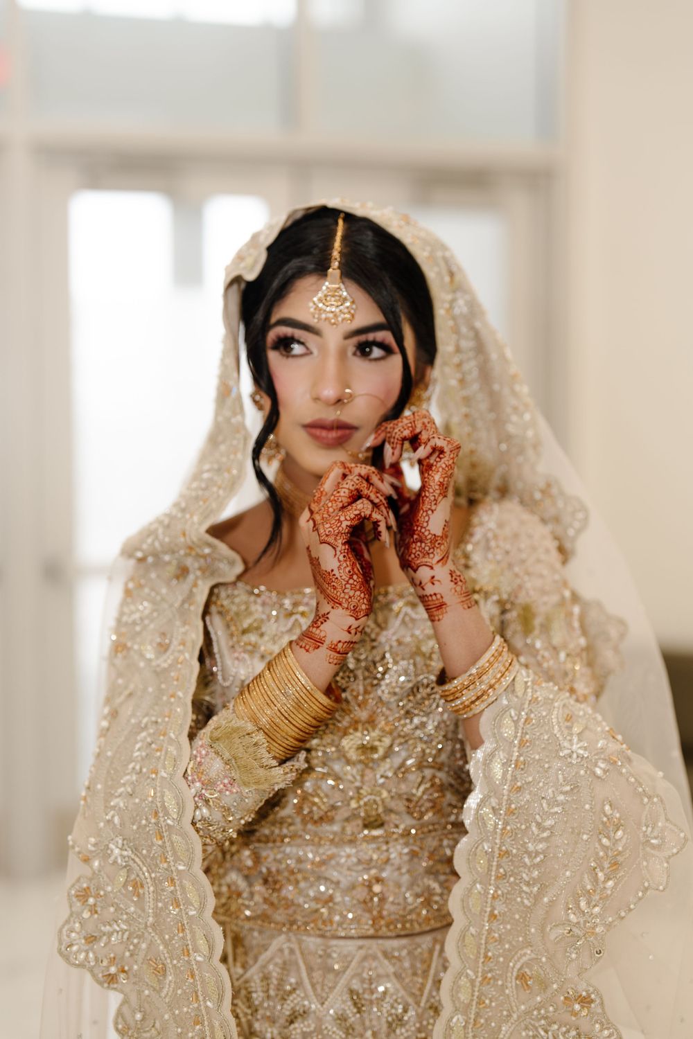 South Asian bride in ivory embroidered lehenga with henna hands, gold maang tikka, and dupatta veil.