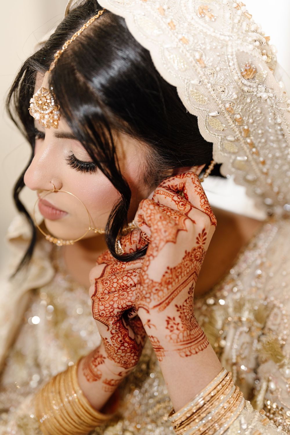 South Asian bride in ivory lehenga putting on gold earring with intricate mehndi hands and maang tikka.