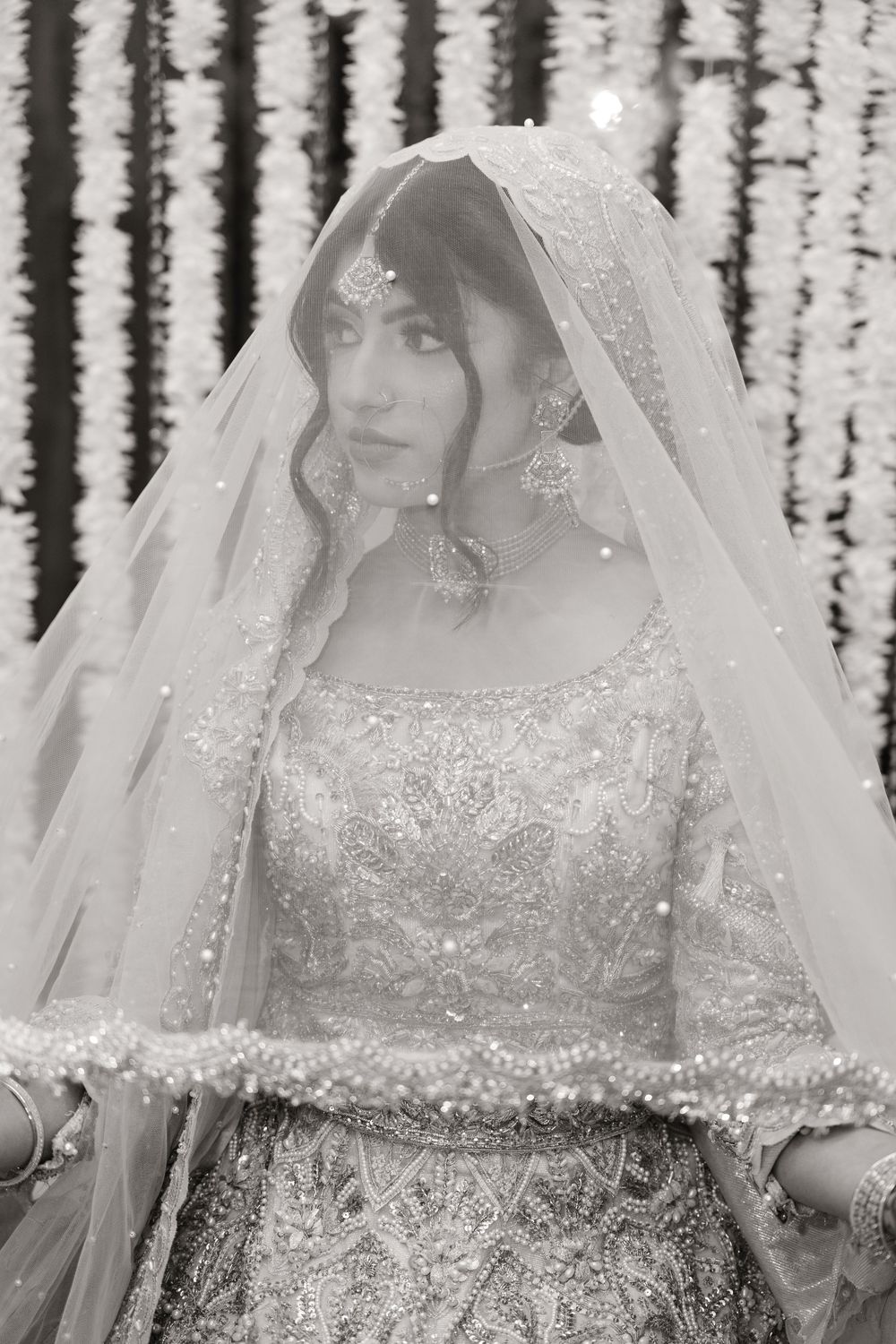Black and white portrait of South Asian bride in embroidered lehenga and veil with floral backdrop.