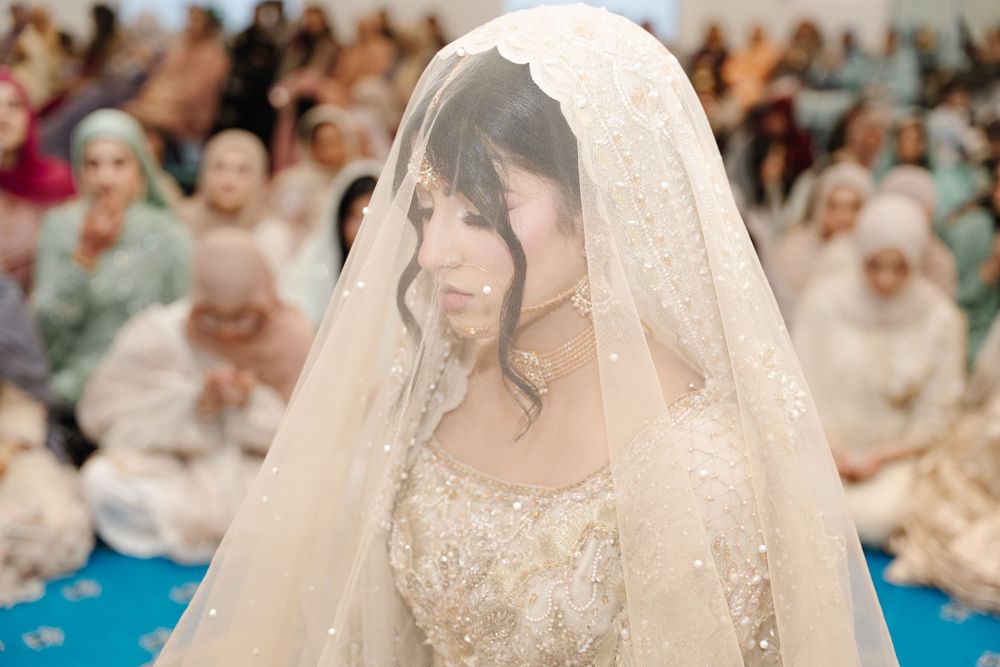 South Asian bride draped in ornate beaded ivory veil sits during traditional wedding ceremony with guests.