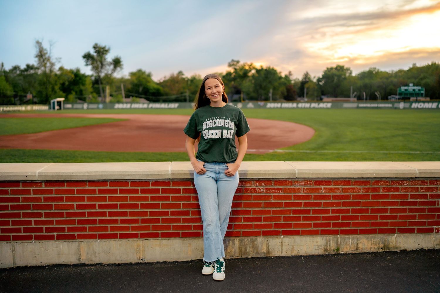 A student in a green university shirt leans against a brick wall at a baseball field during sunset.
