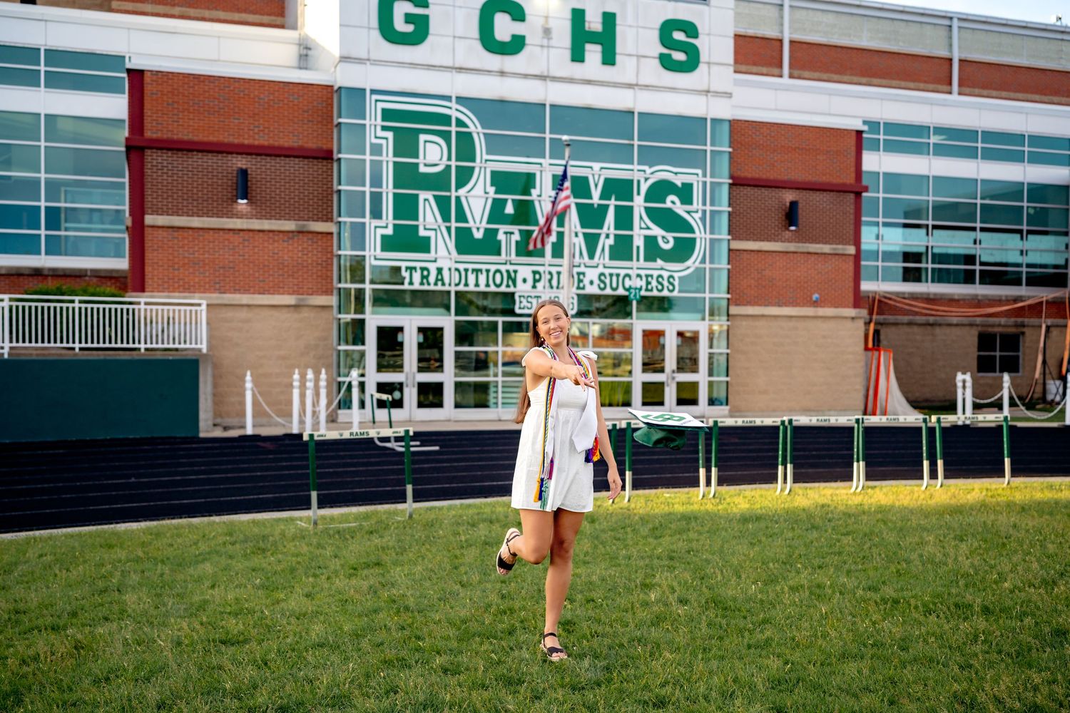 A person in a white dress stands on grass in front of a school building with green GCHS RAMS signage.