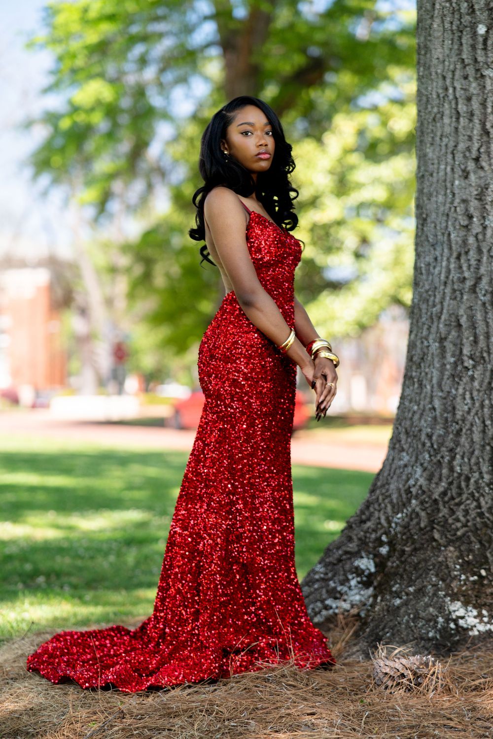A person in a sparkling red evening gown poses gracefully among trees in an outdoor setting.
